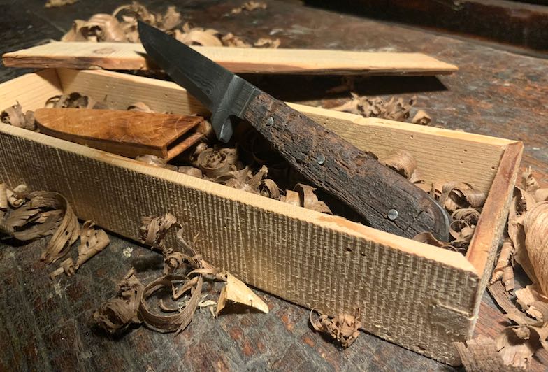 Damastmesser mit rustikalem Griff aus altem Eichenholz und Kirsch-Scheide in Holz-Box mit Hobelspänen auf Werkbank