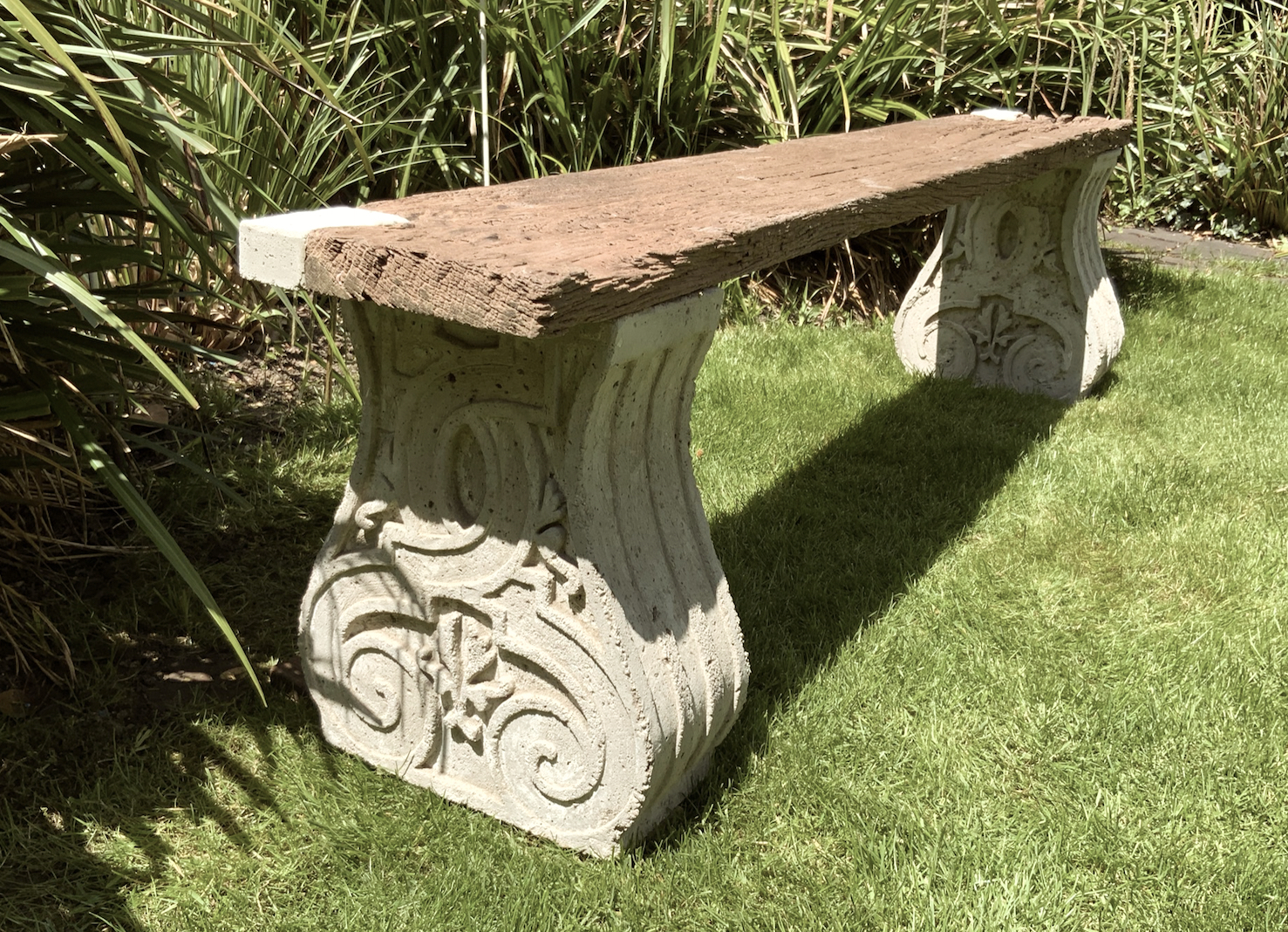 Sitzbank aus altem, verwittertem Holz auf mit historischen Ornamenten verzierten Betonsockel im Garten