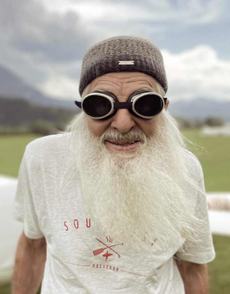90jähriger Manfred Fritz Threimer mit Fliegerbrille, weißem Bart, langen Haaren und Stirnband vor Wiese